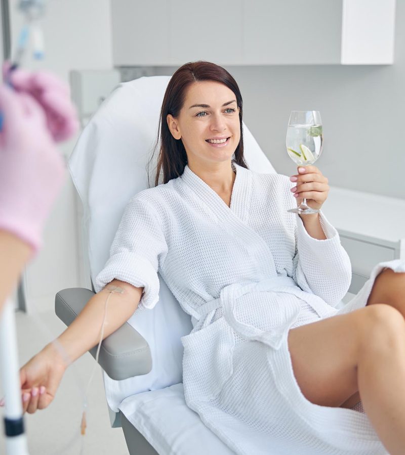 Joyful female patient undergoing the intravenous therapy Smiling good-looking dark-haired spa client relaxing with a glass of beverage in a cosmetic chair